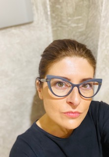 Donna con occhiali blu e capelli raccolti, posa davanti a parete chiara, espressione pensierosa.